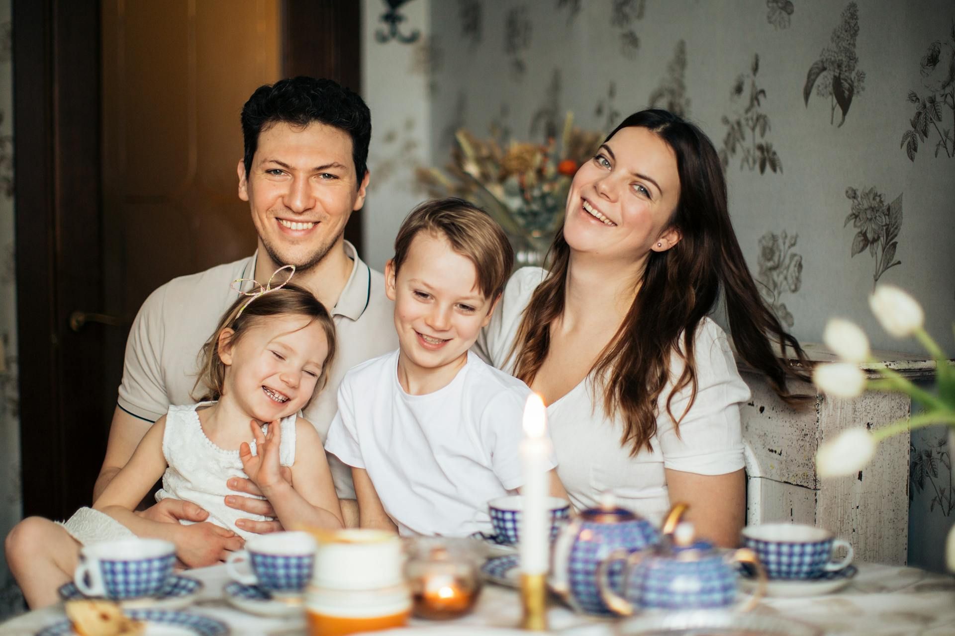 A happy family of four sitting together at a table for tea, smiling warmly in a softly lit, cozy indoor setting.