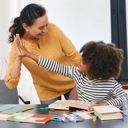 A teacher and student high-five while sitting at a desk with books and school supplies.