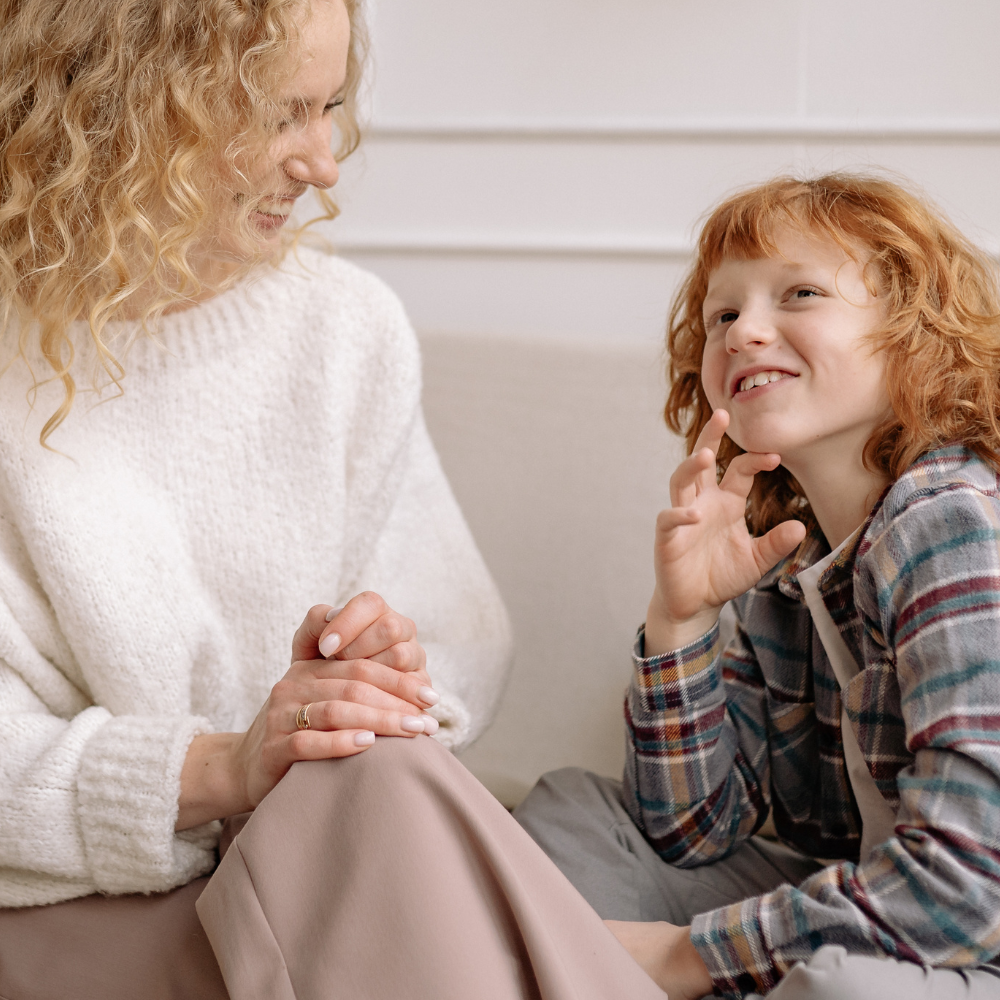 A curly-haired adult in a white sweater sits with a red-headed child in a plaid shirt, both smiling during a conversation.