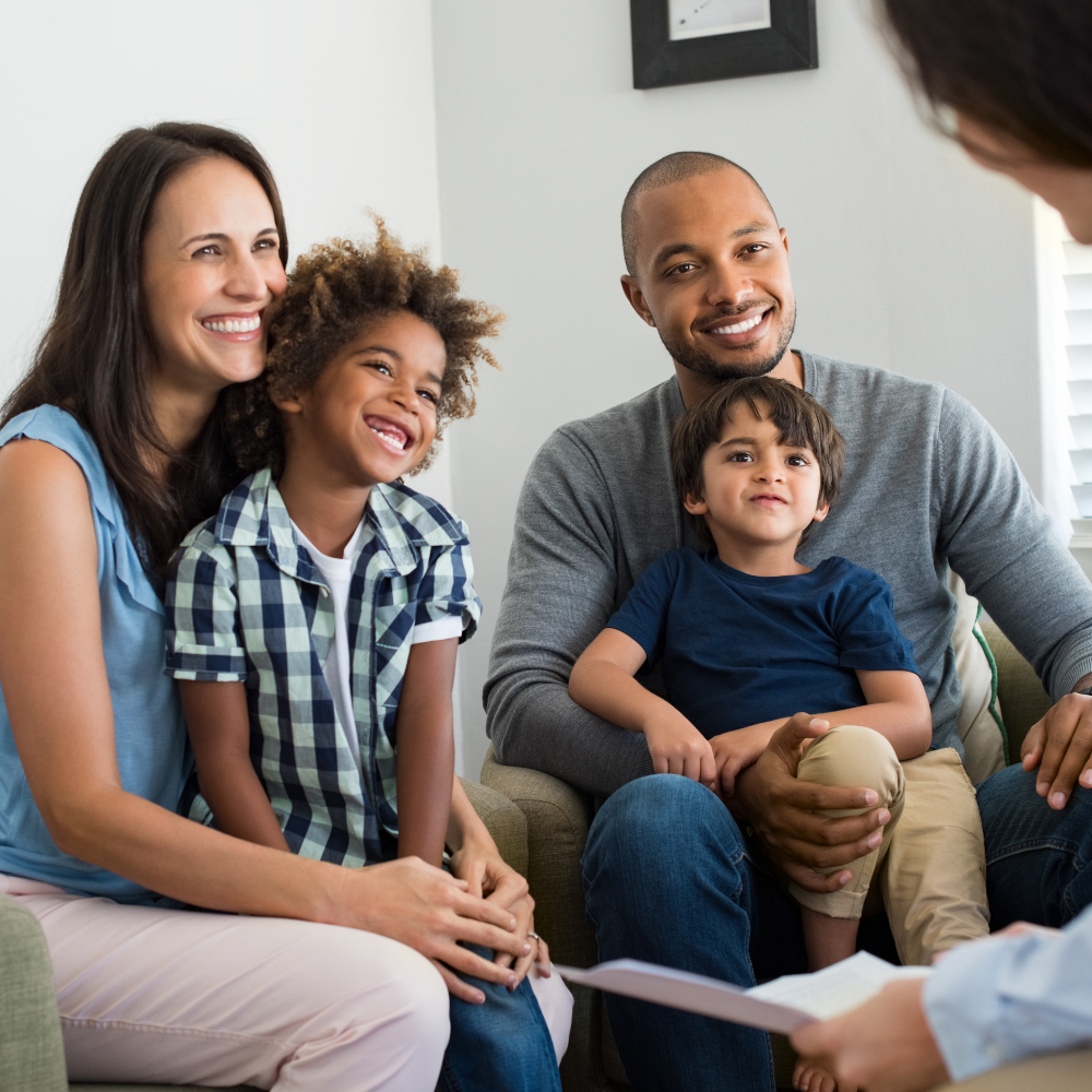A family smiling while sitting together and talking with someone off-screen in an office or counseling setting.
