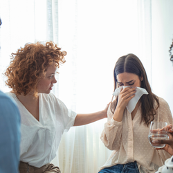 A person comforts a distressed individual covering their face with a tissue, with a glass of water held nearby.