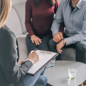 A therapist holds a clipboard while meeting with a couple sitting on a sofa in an office setting.