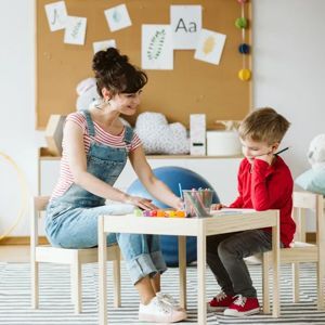 A teacher and a child sit at a small wooden table, drawing together in a bright, colorful classroom.