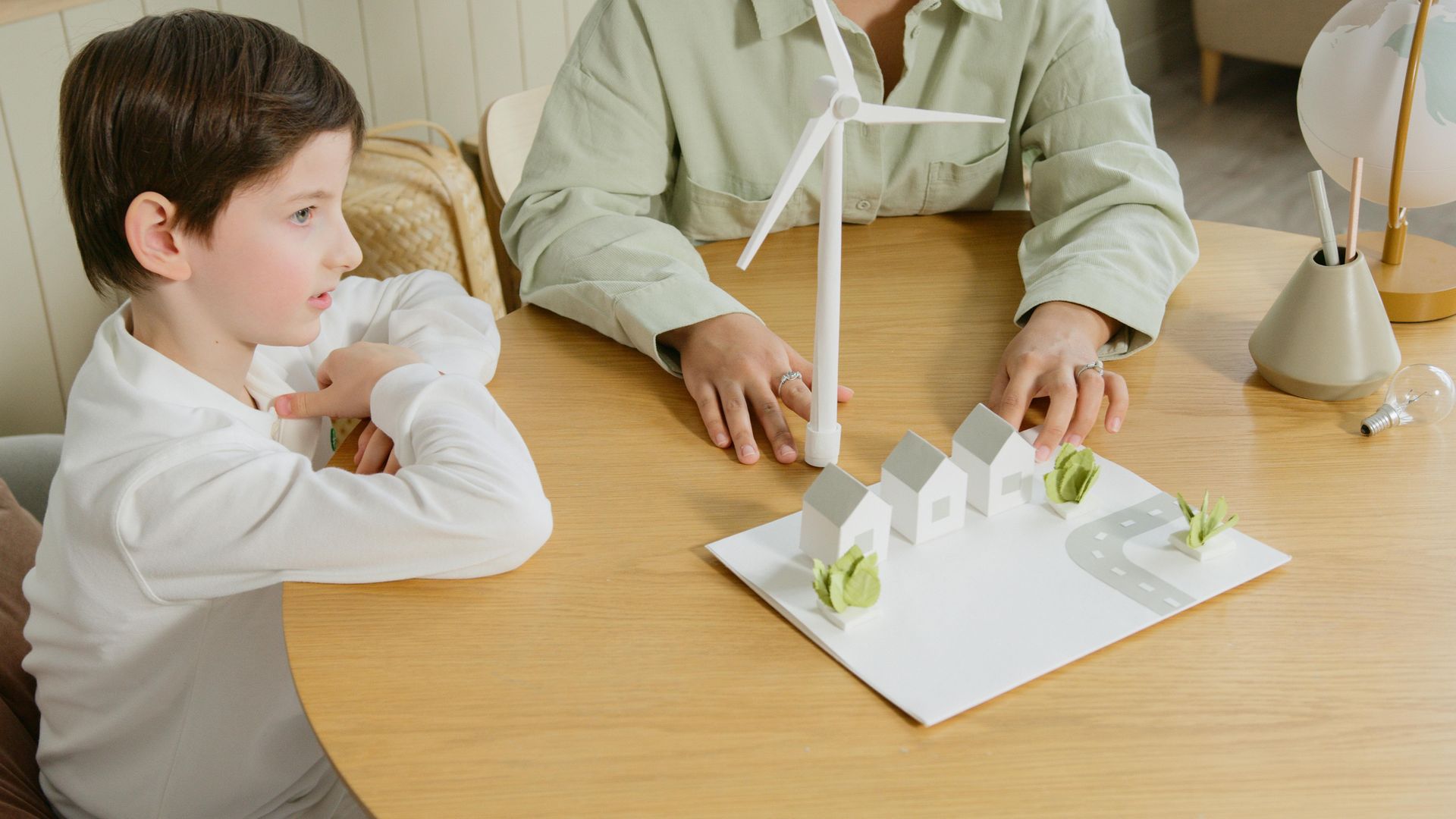 A child sits at a round wooden table with an adult, building a model of houses and a white wind turbine.