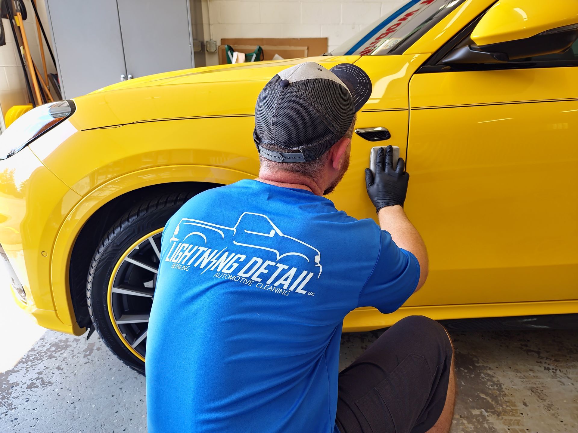 Man applying product to yellow car door; wearing black gloves, blue Lightning Detail shirt.