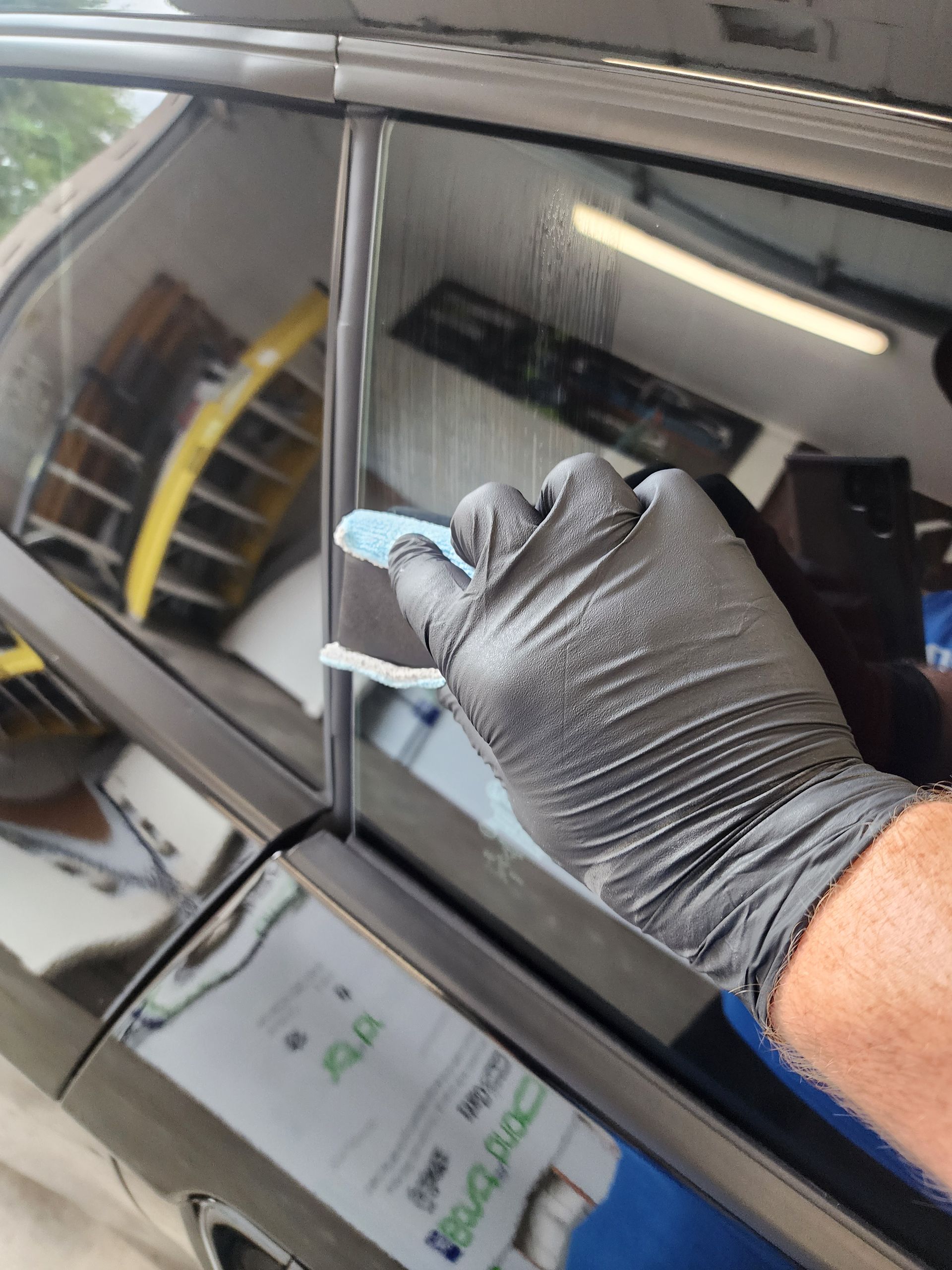 A gloved hand cleans a car window's track. Black car, shiny glass, indoor garage setting.