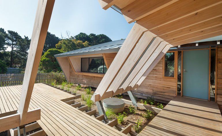 Wooden house with angled beams and a deck, incorporating landscaping and a blue door. — On The Rise Builders in Maroochydore, QLD