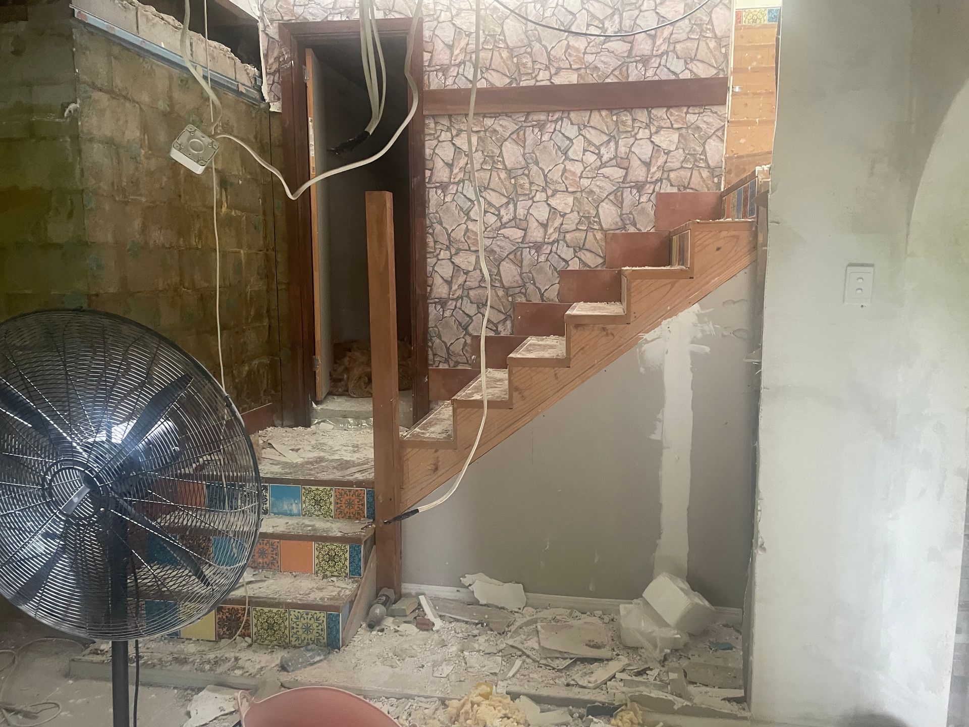 Renovated interior with a wooden staircase, debris, and a large fan in the foreground.