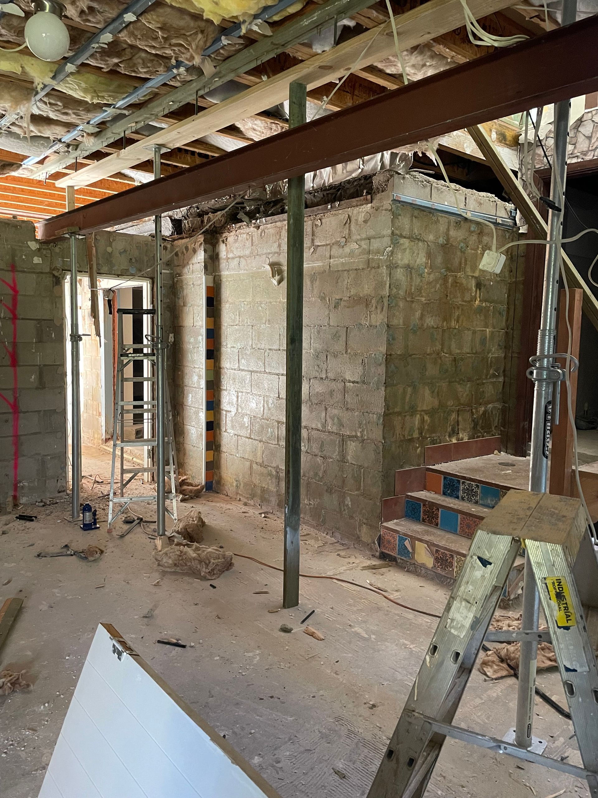 Interior renovation, a steel beam supported by vertical jacks over a brick wall, ladder nearby.