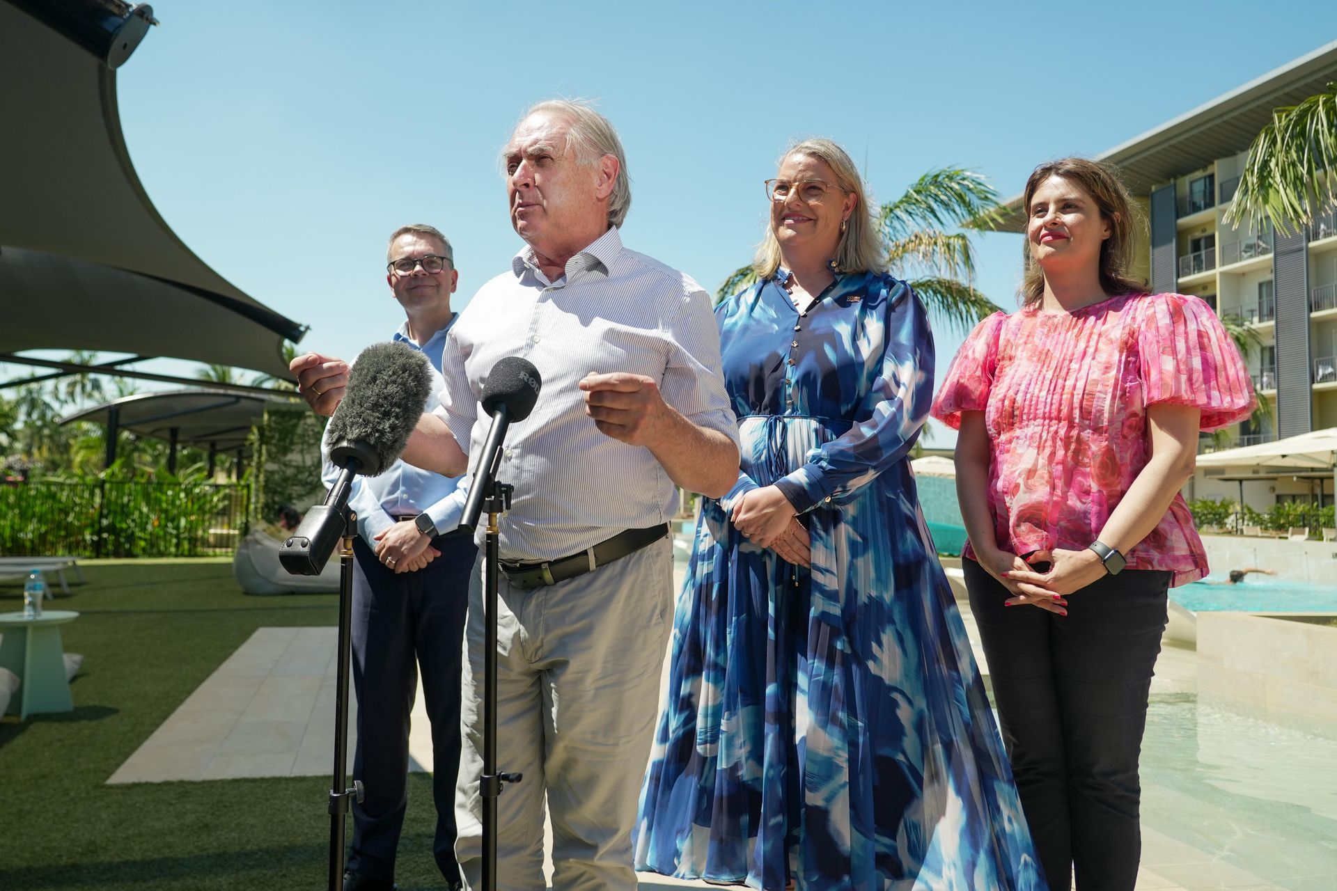 Four people outdoors, one speaking into microphones while holding papers; building and palm trees in background.