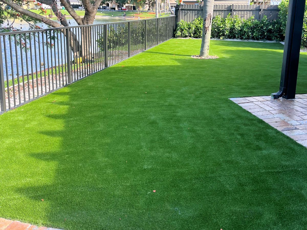 Green artificial grass backyard with black fence, trees, and small lake.