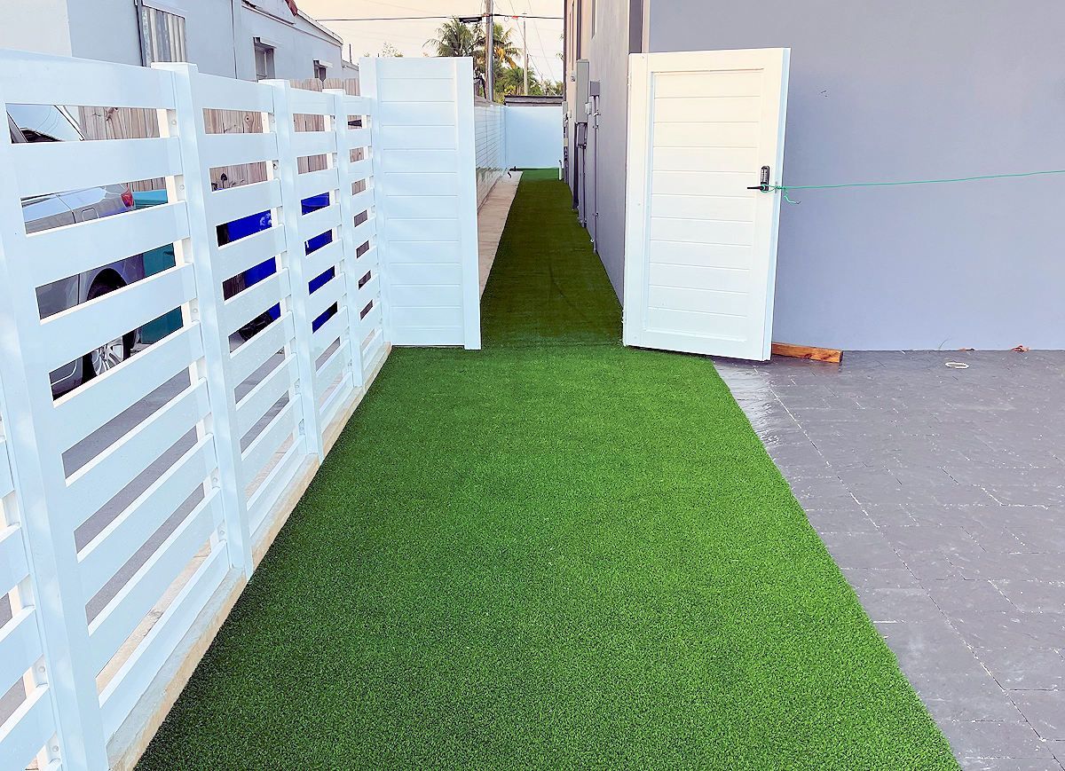 Narrow strip of green turf between white fences and a door. Concrete patio.