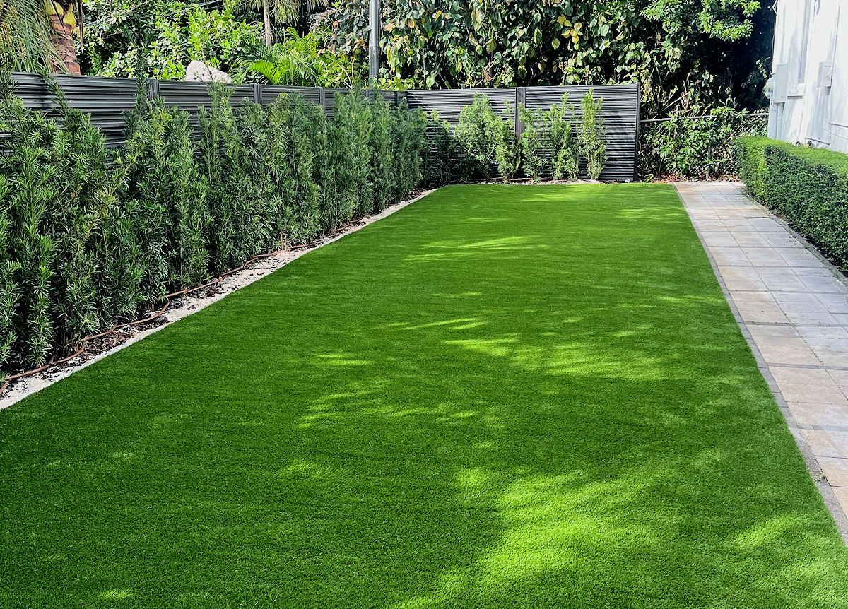 Green artificial turf lawn bordered by hedges, gravel, and a stone path.