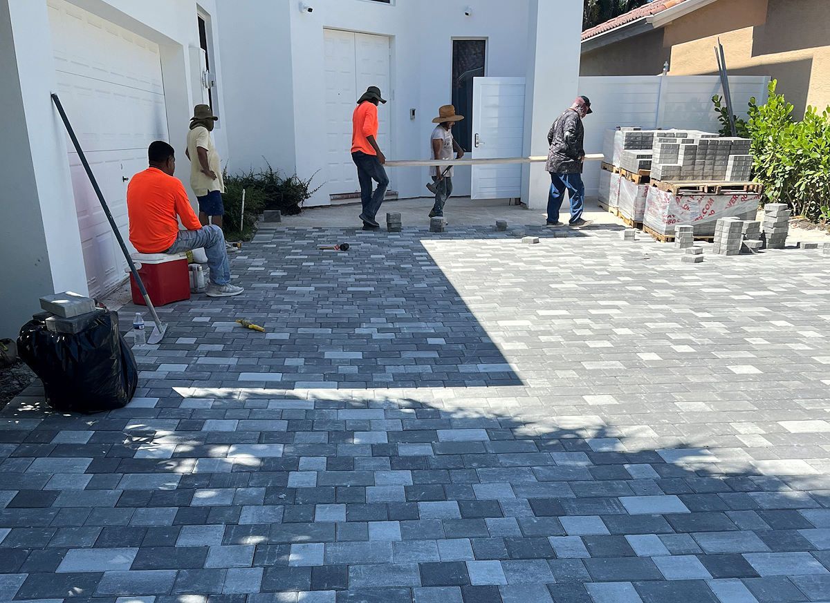 Workers laying pavers in a driveway; gray and blue brick pattern, building in background, sunny day.