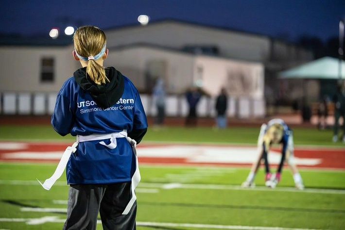 Girl in blue sports jersey watches a flag football game on a lit field at night.