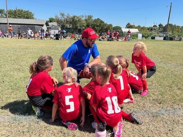 Youth soccer coach kneels with red-uniformed players on a grassy field during a game break