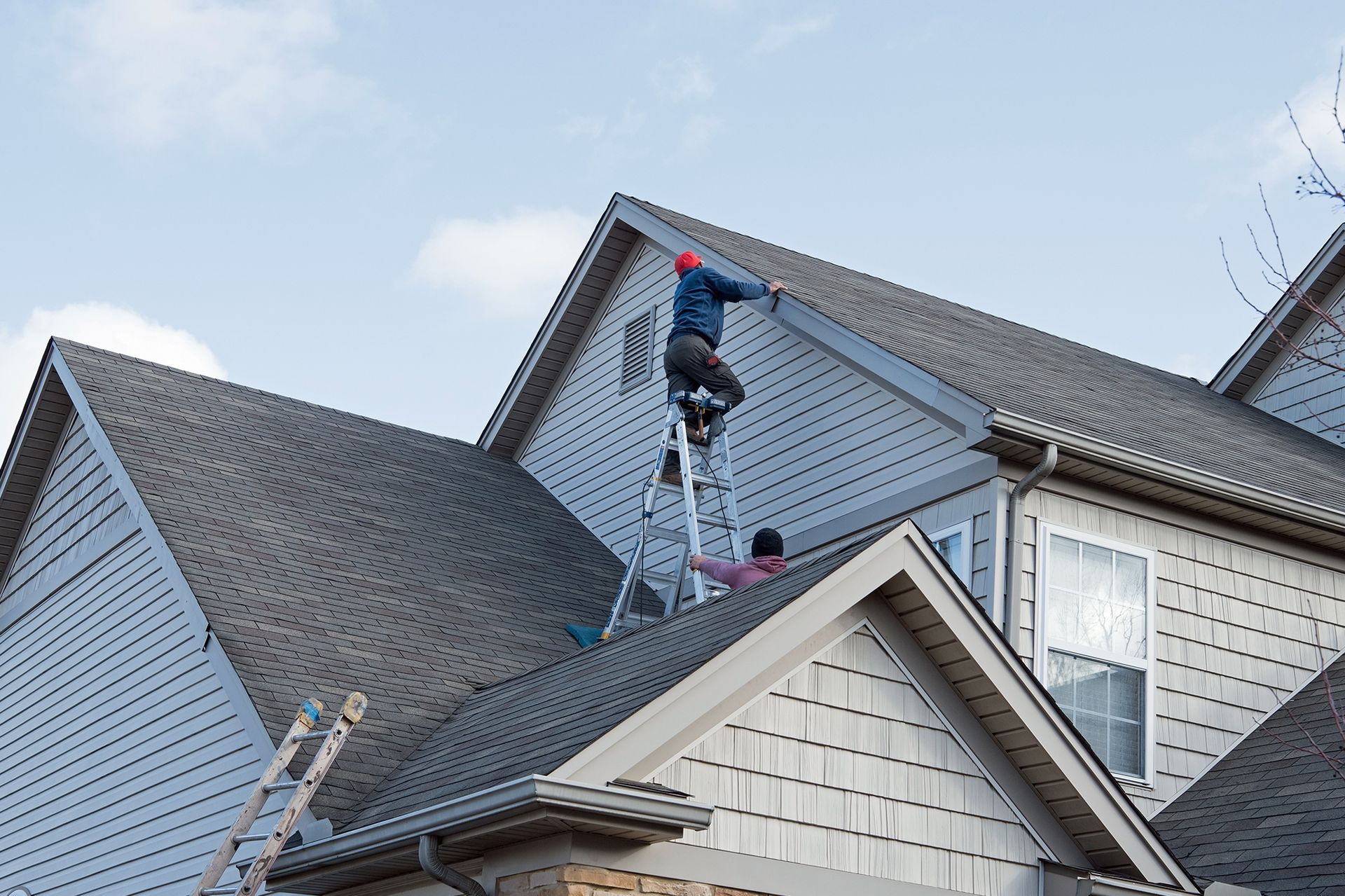 Two people on a roof, one on a ladder. House exterior, overcast sky.