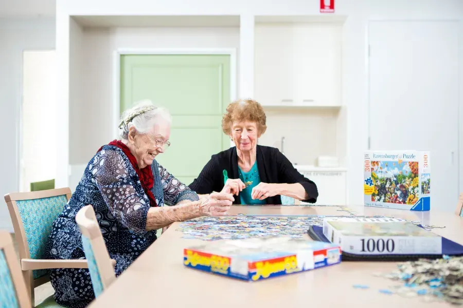 Two elderly women are at Beauaraba Living, they are sitting at a table playing a puzzle.