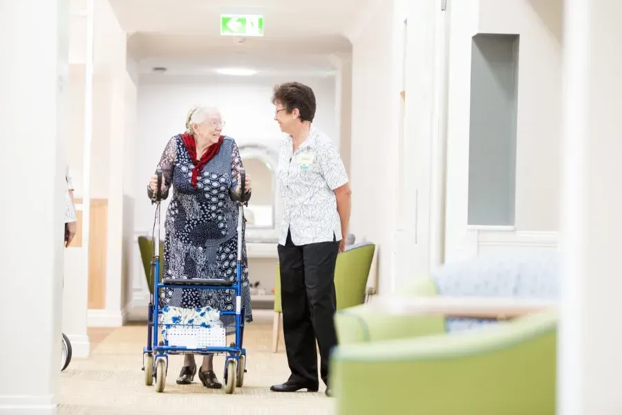 A nurse is helping an elderly woman with a walker in a hallway at Beauaraba Living.