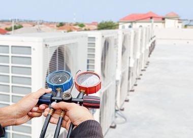 Hands holding blue and red manifold gauges beside rooftop air-conditioning units