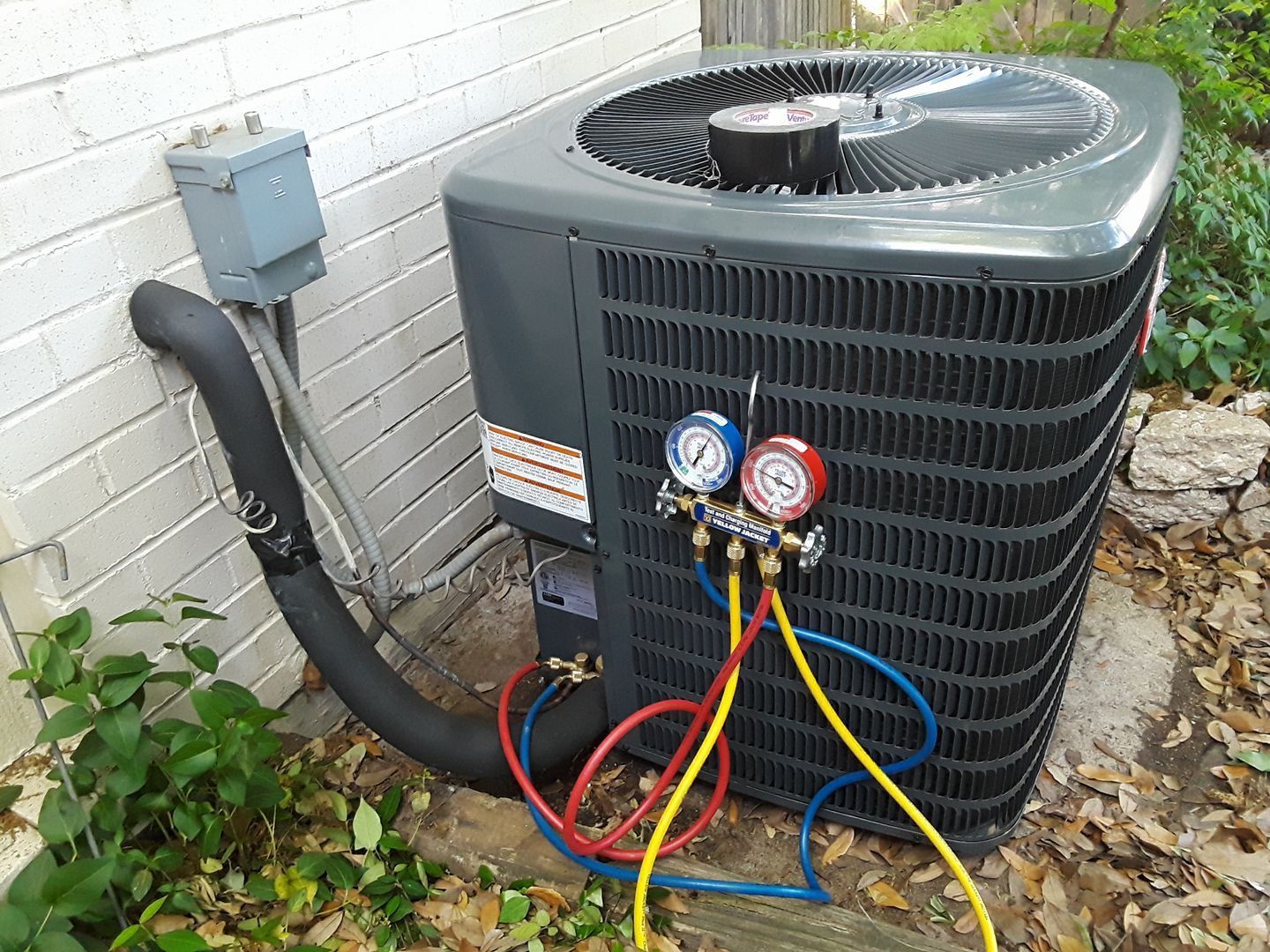 Outdoor air conditioner unit with exposed gauges and colored hoses beside a brick wall