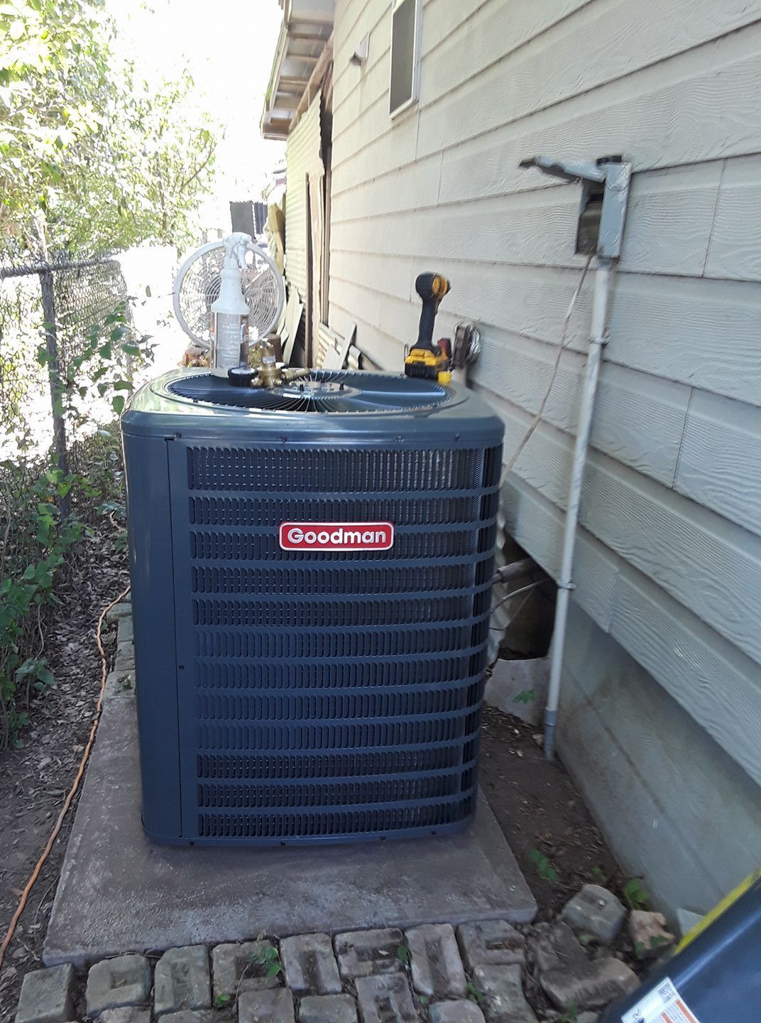 Outdoor Goodman air conditioner unit beside a house wall with a service gauge on top
