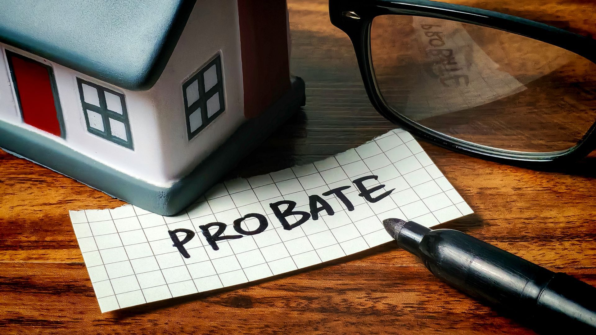 Note labeled probate next to a model house, pen, and glasses on a wooden desk