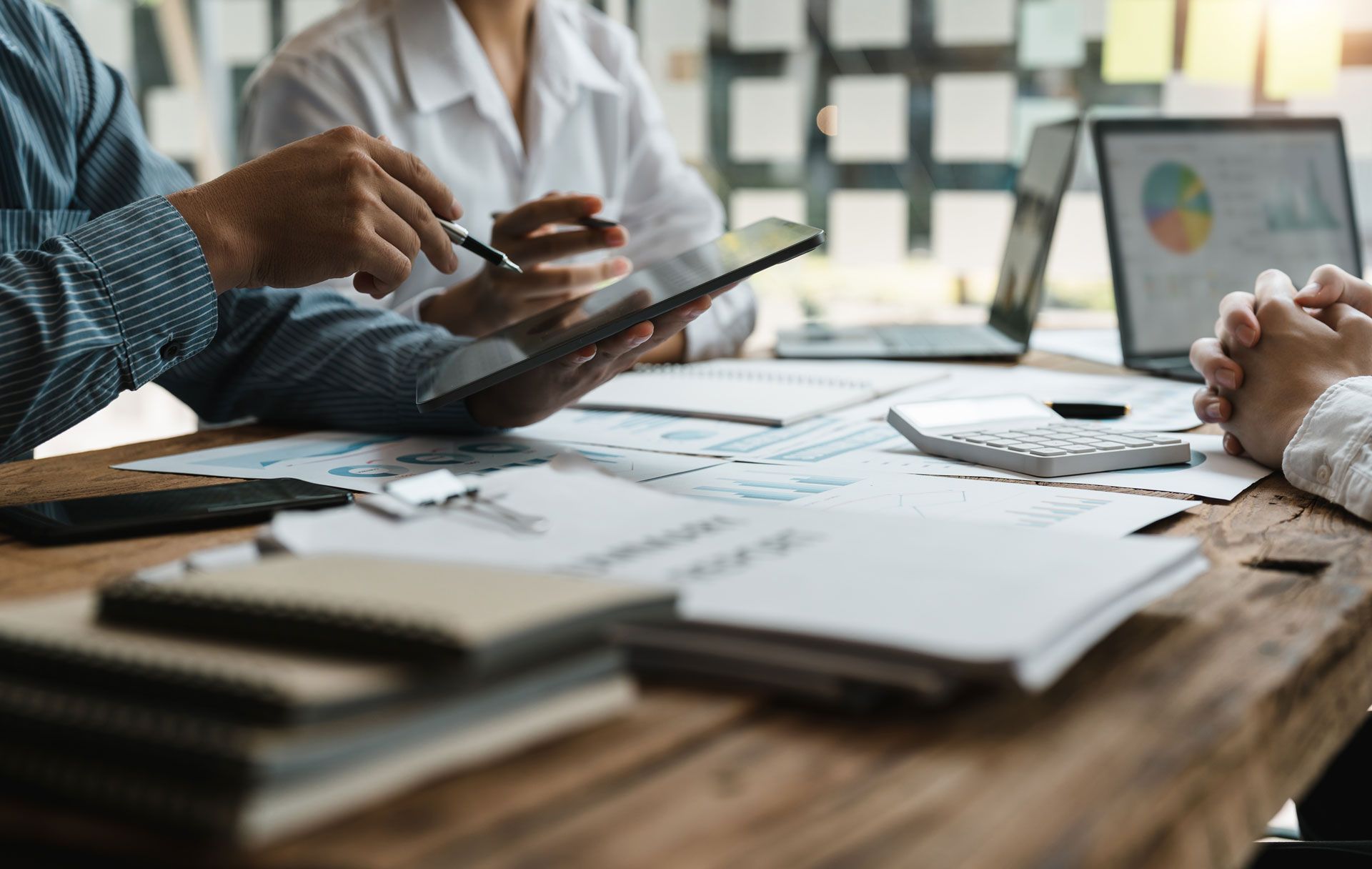 People in a meeting review documents and a tablet. A laptop with a pie chart is also on the table.