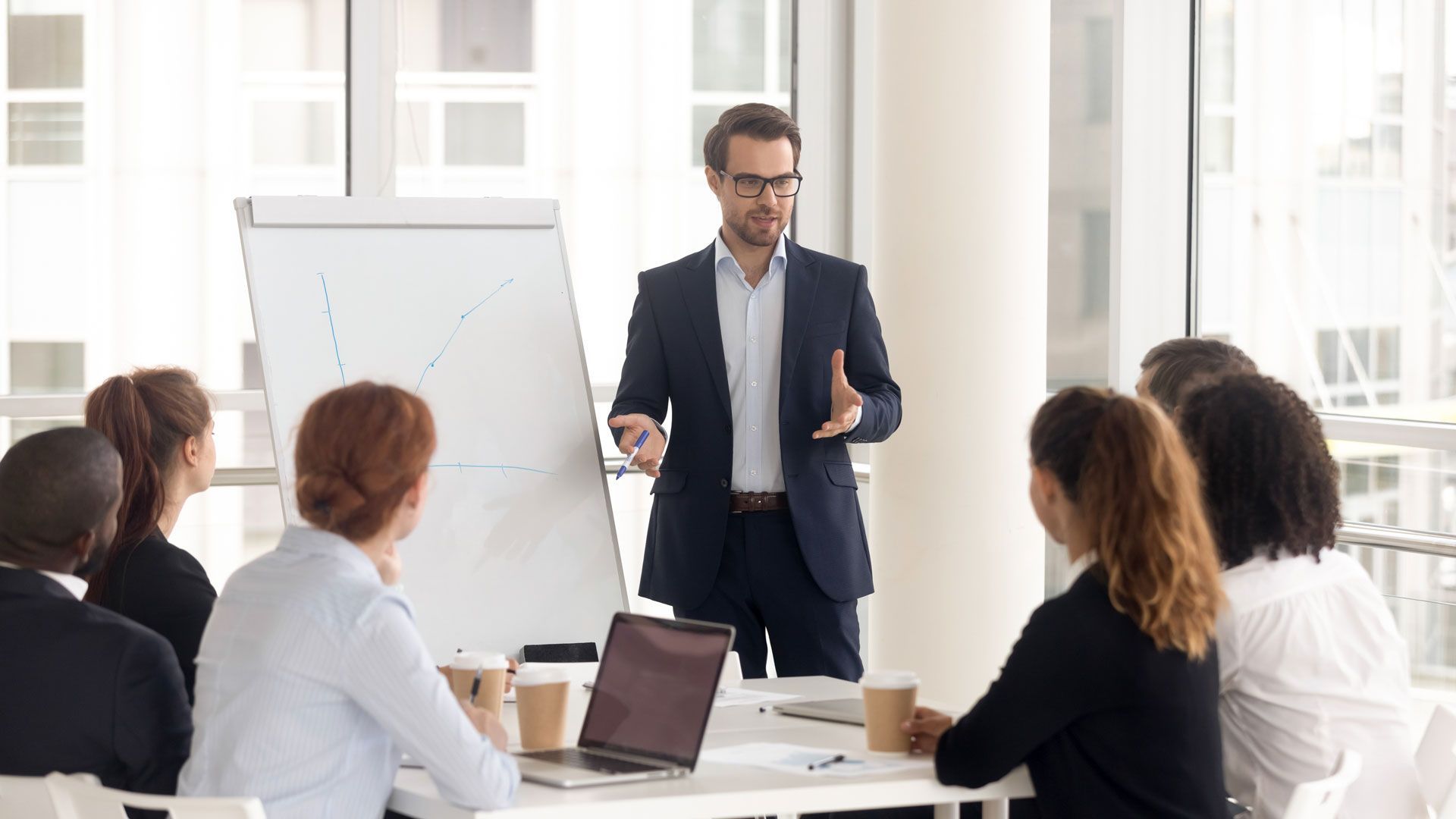 A man in a suit presents to a group of colleagues at a table in an office. He gestures toward a flip chart.