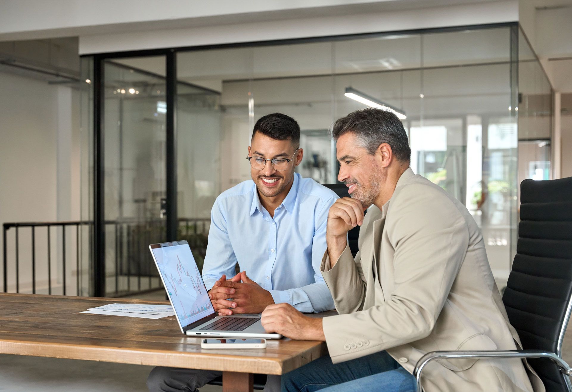 Two men in office setting, looking at laptop screen with smiles. One man in a light blue shirt, the other in a tan jacket.