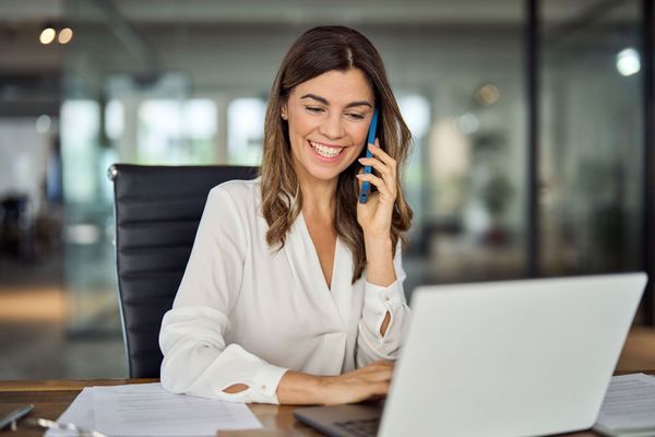Woman in a white blouse smiles while talking on the phone and working on a laptop in an office.