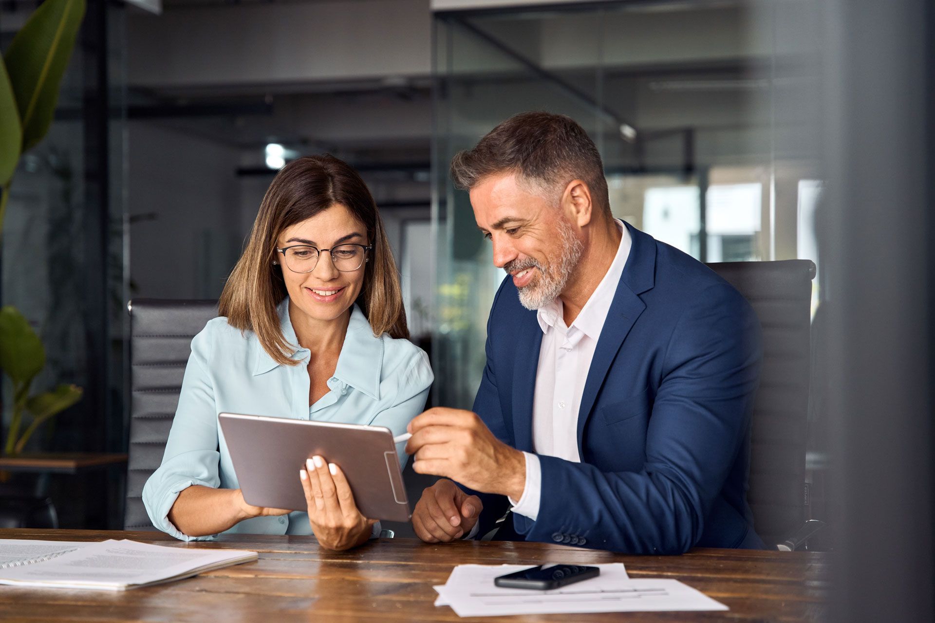 Woman and man looking at a tablet in an office. The woman wears glasses and a light blue shirt. The man wears a blue suit.