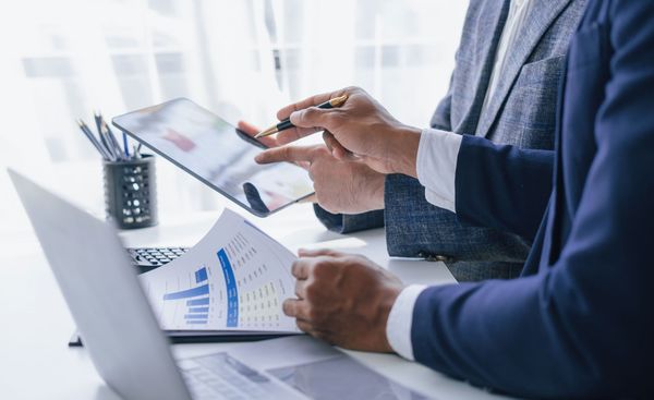 Two businesspeople in suits reviewing financial documents and a tablet at a desk.