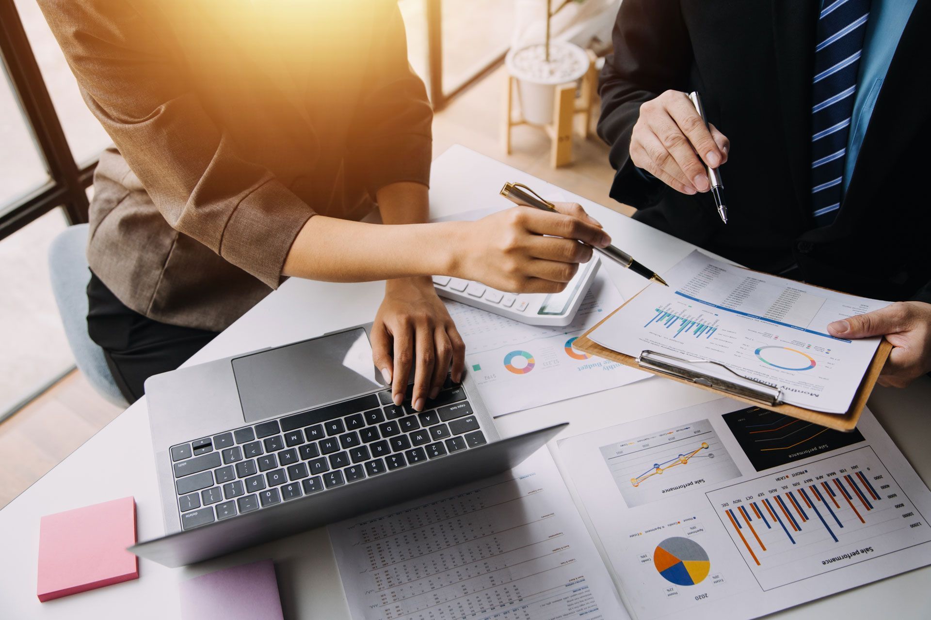 Two people reviewing financial data at a desk with a laptop, charts, and calculator. One points at a document.