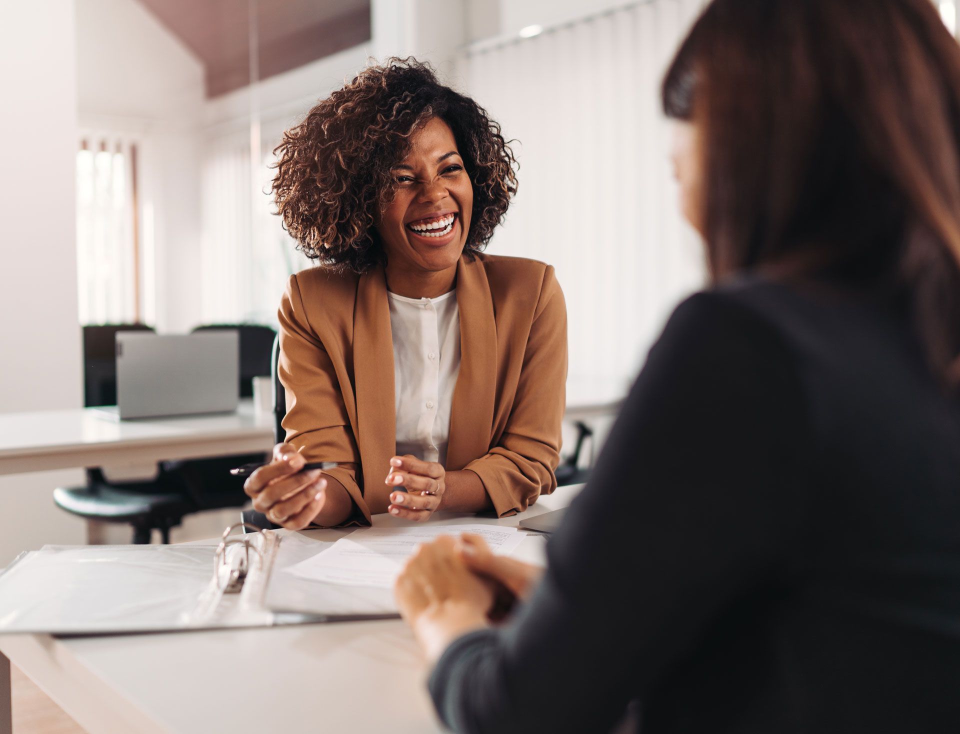 Woman in tan blazer smiles and gestures while speaking to another woman in a business setting.