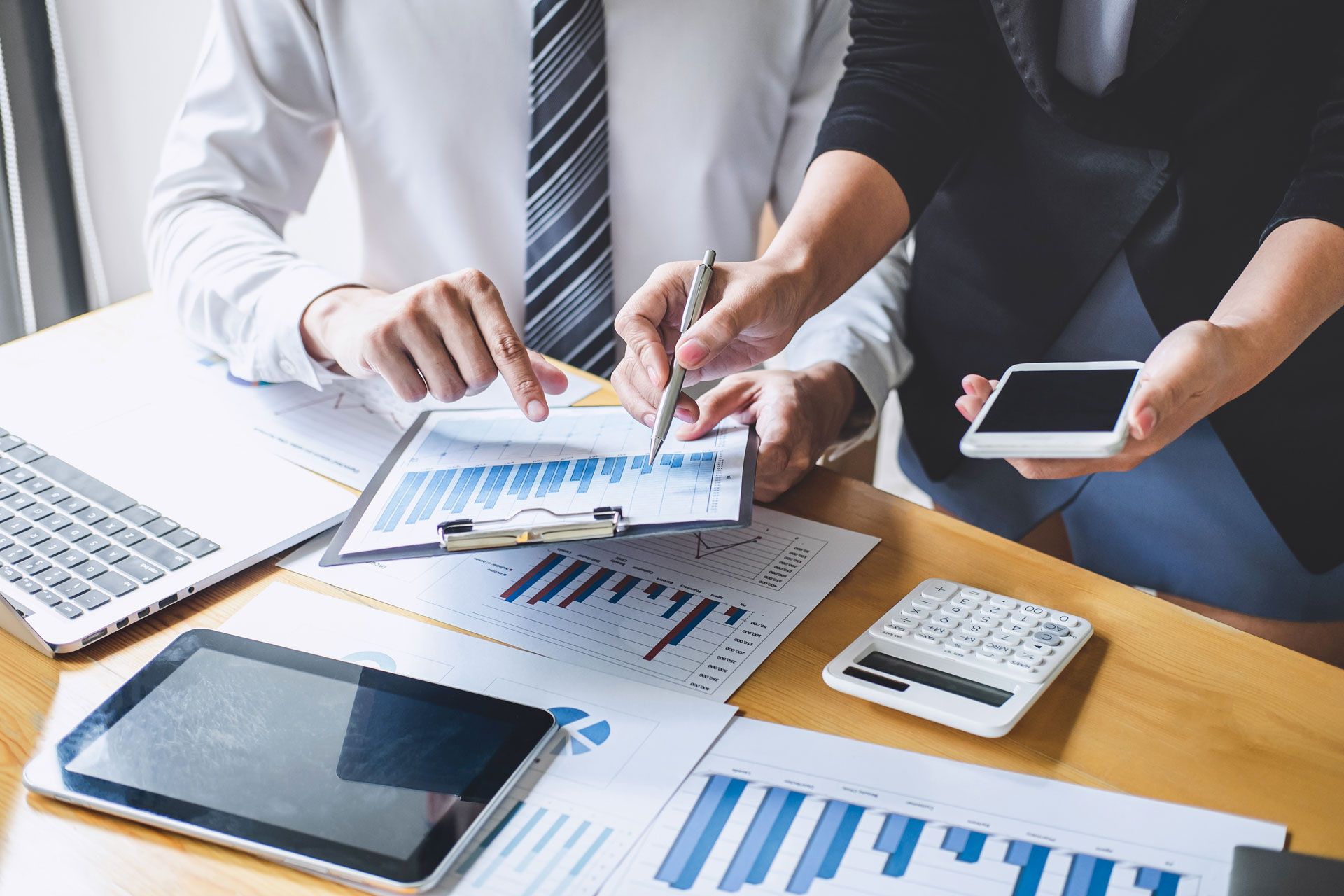 Two people reviewing financial charts at a desk, pointing to the data, with a laptop, tablet, calculator, and phone nearby.