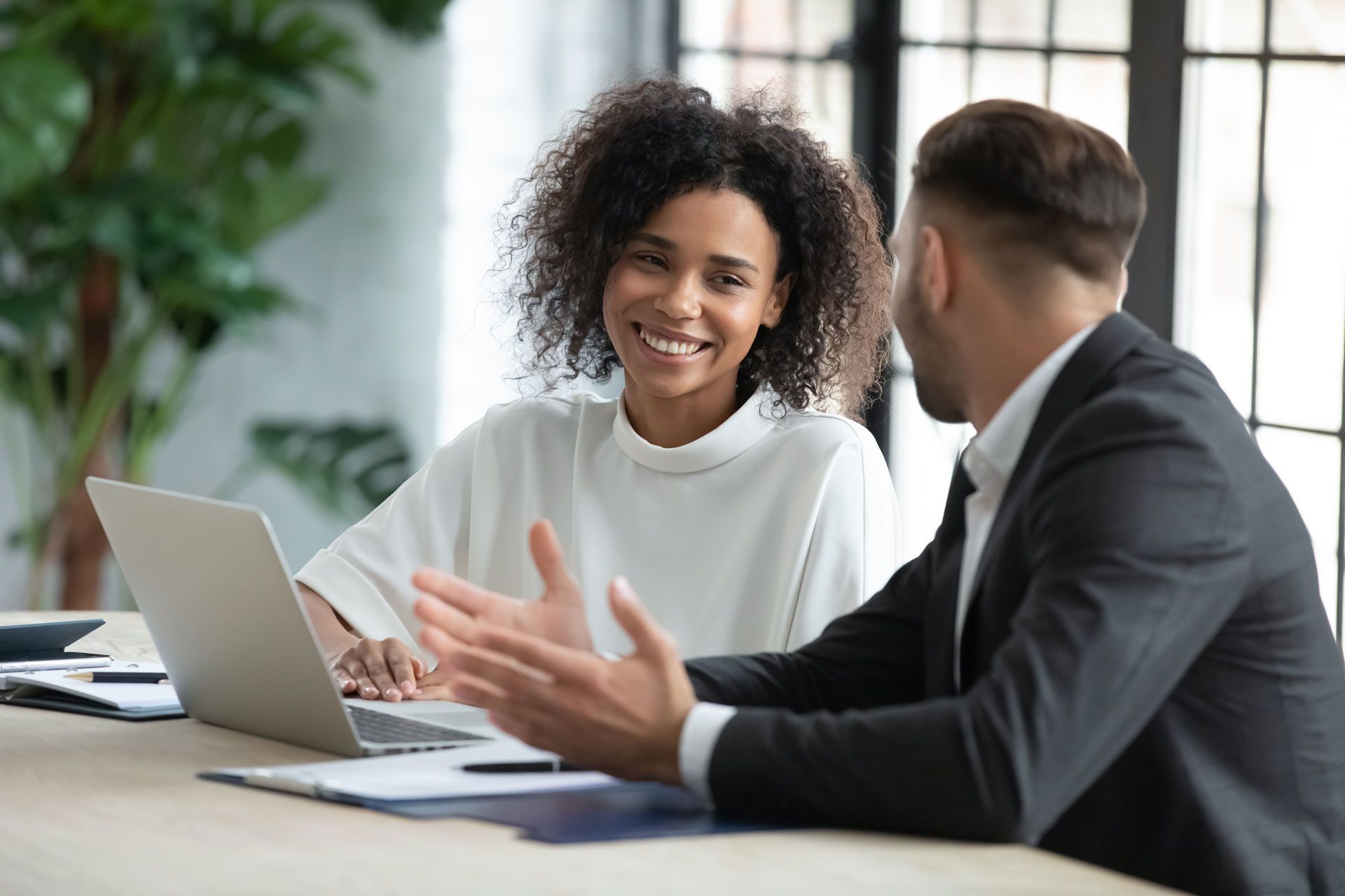 A Black woman smiles at a man, both seated at a table with a laptop. They appear to be collaborating in an office setting.