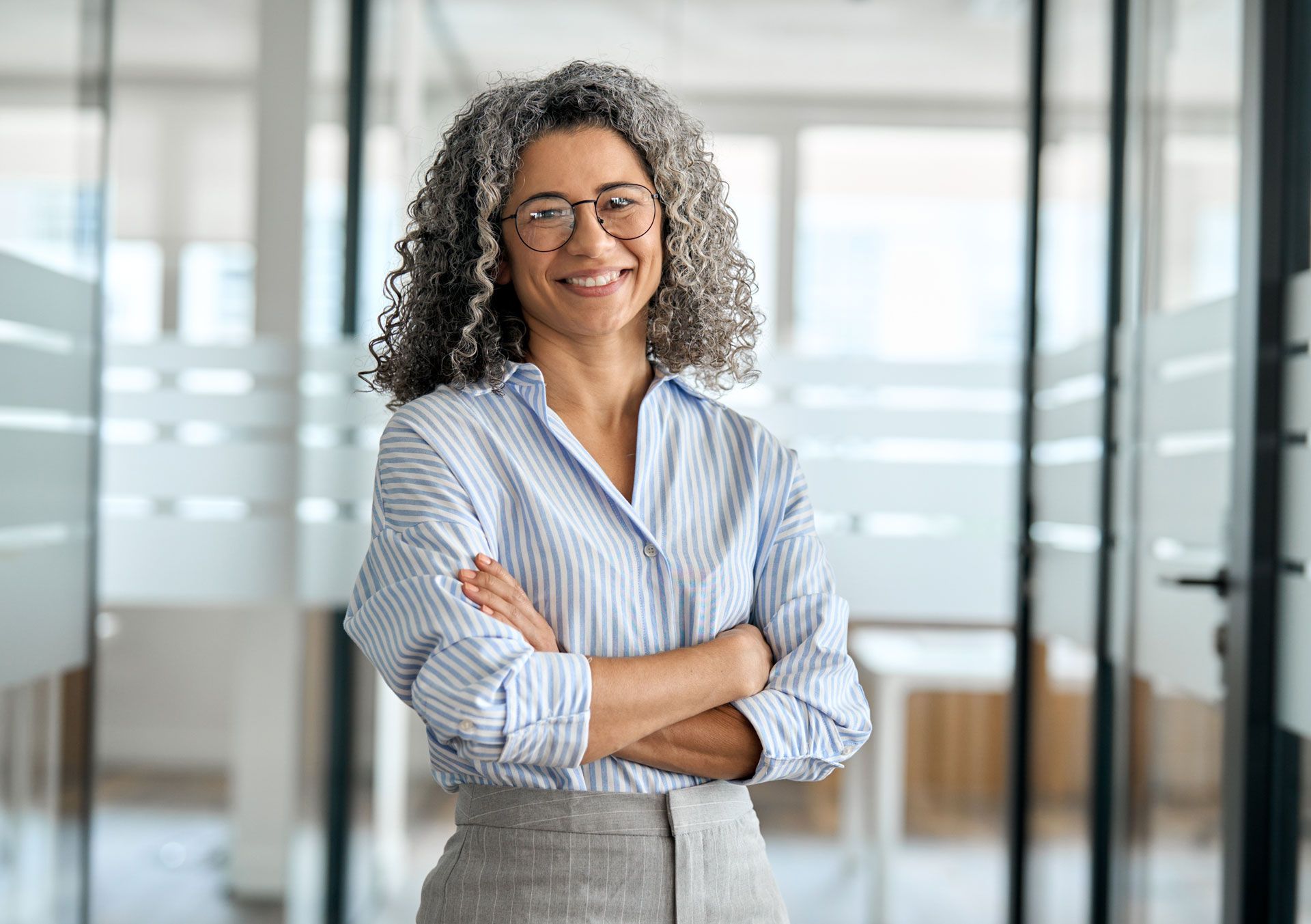 Woman with gray curly hair, wearing glasses and a striped shirt, smiling with arms crossed in an office hallway.