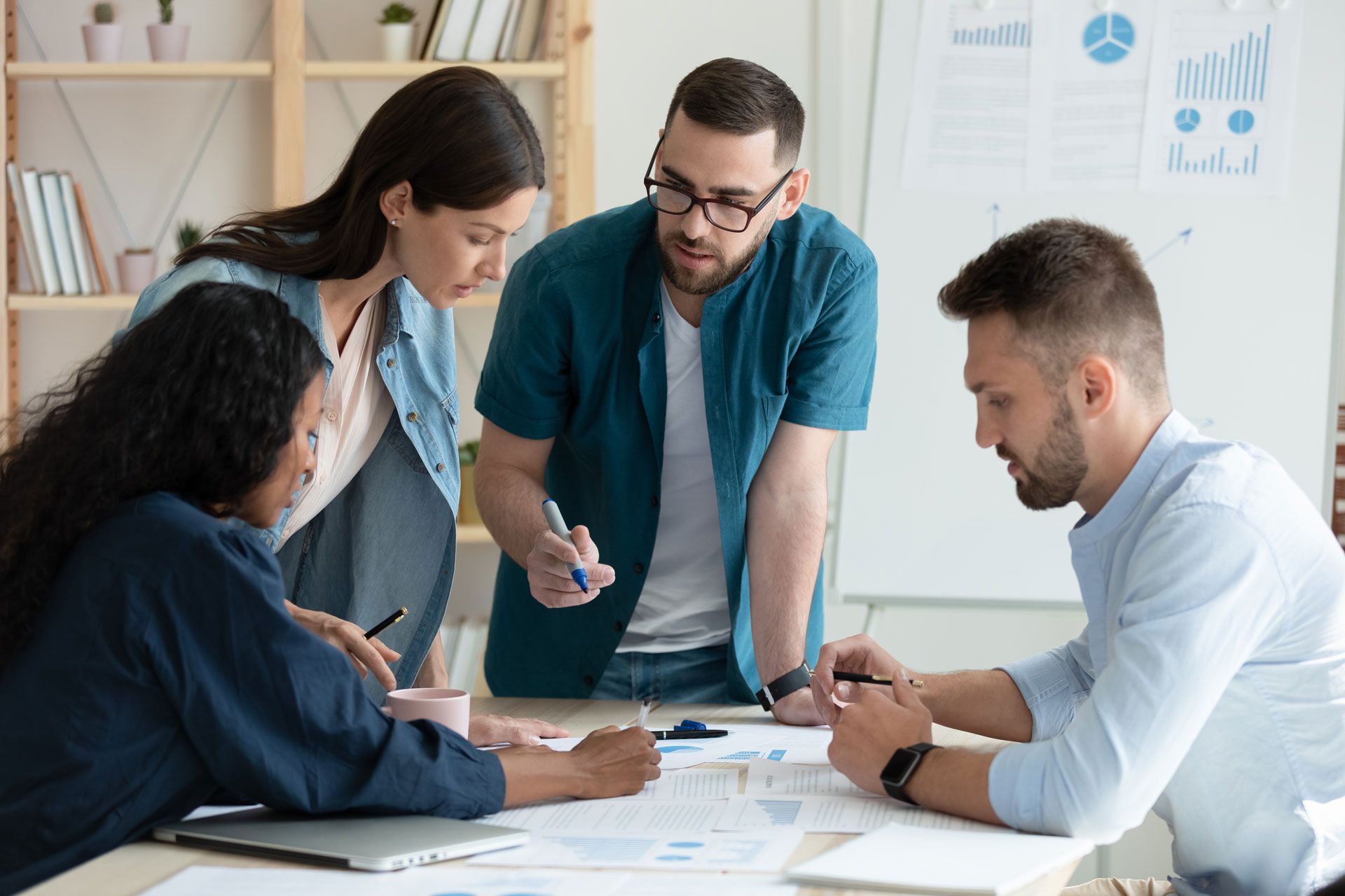 Four colleagues collaborating at a table, focused on papers, one pointing with a pen, whiteboard with charts in background.