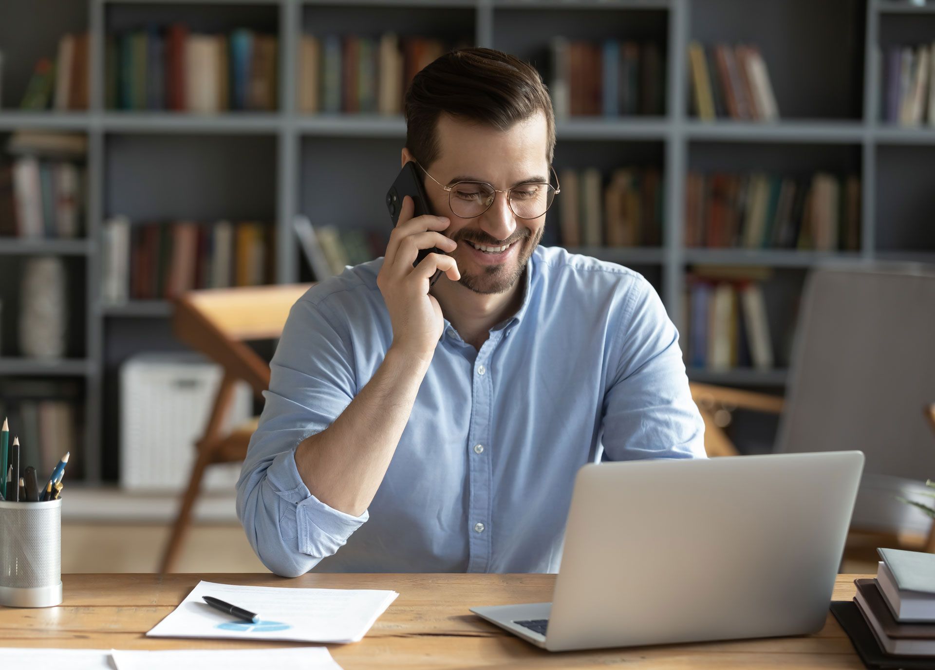 Man in blue shirt smiling while talking on a cell phone, seated at a desk with a laptop, papers, and books.