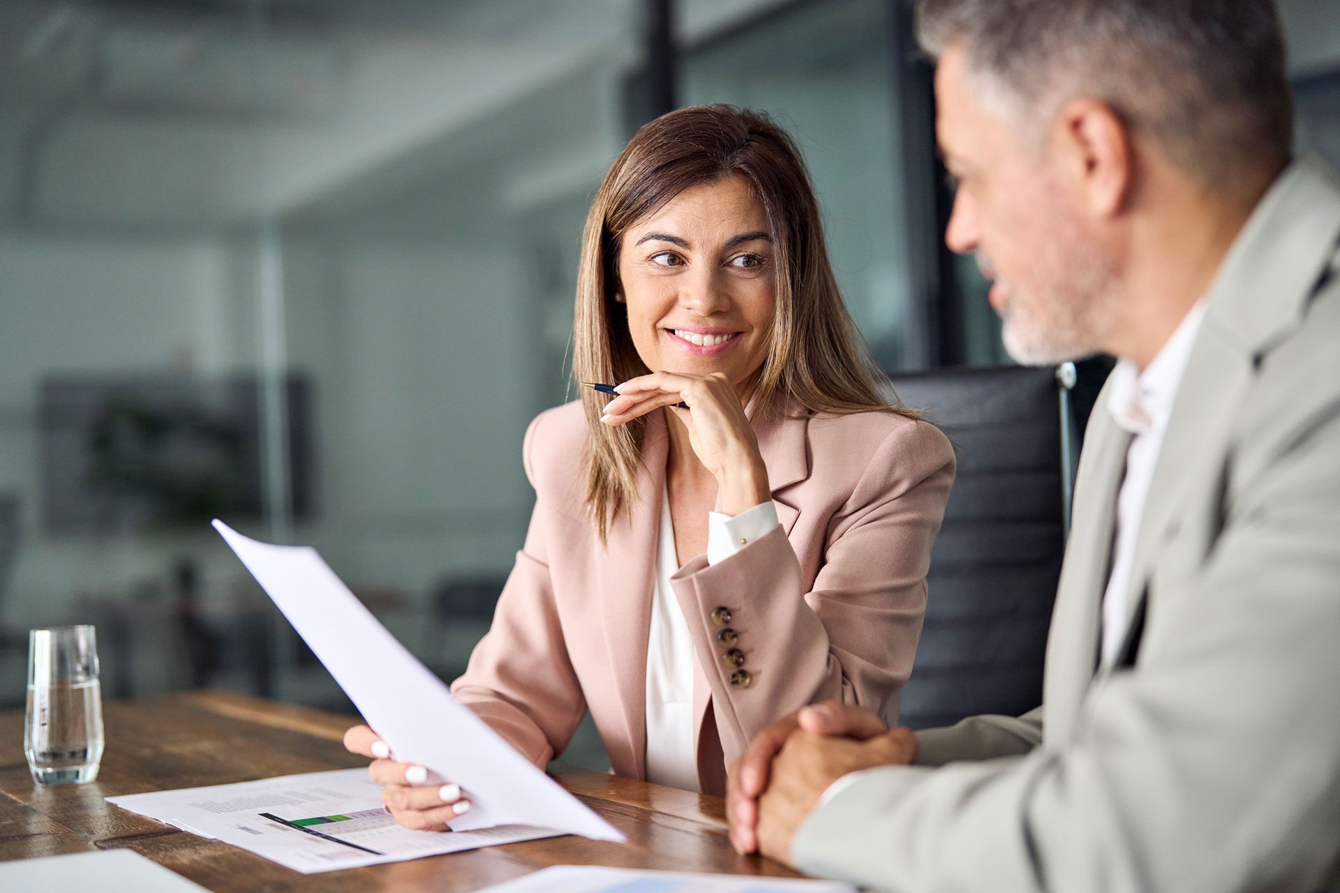 Woman in pink blazer smiling, reviewing papers with a man in an office setting.