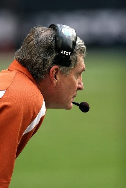 A determined coach stands on an athletic field holding a clipboard, with translucent overlays of a shield, legal gavel, and NCAA logos in the background symbolizing protection and compliance with NCAA rules.