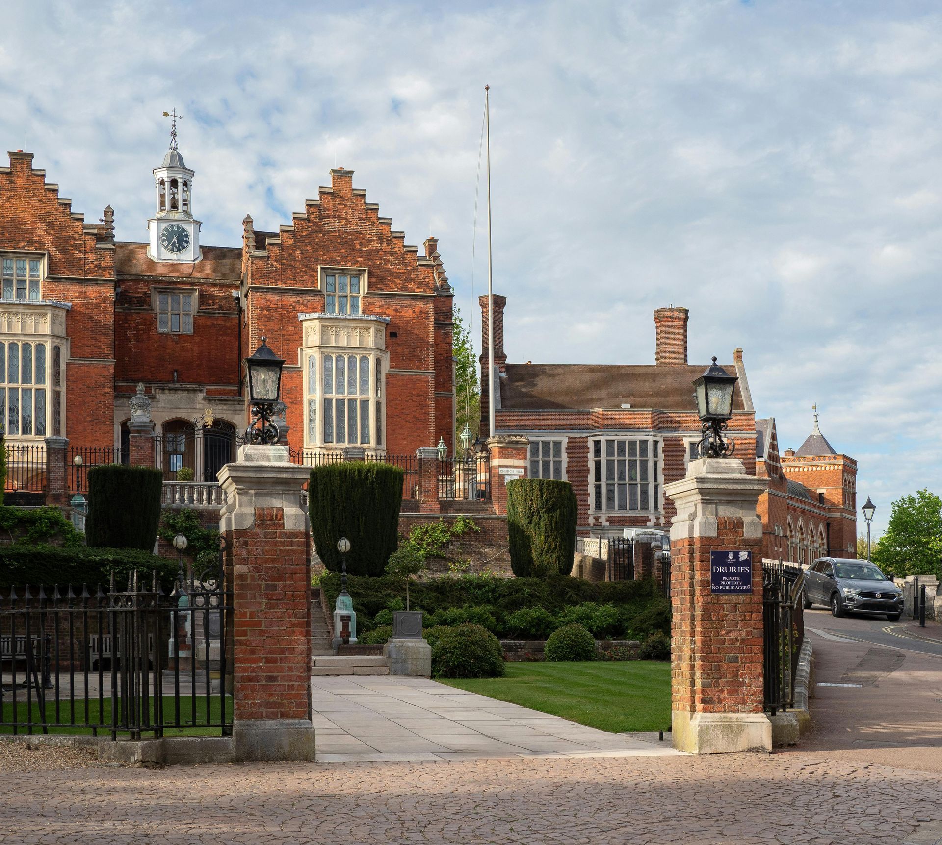 Brick Tudor-style building with clock tower, viewed through open wrought iron gates. Formal garden and driveway in foreground.