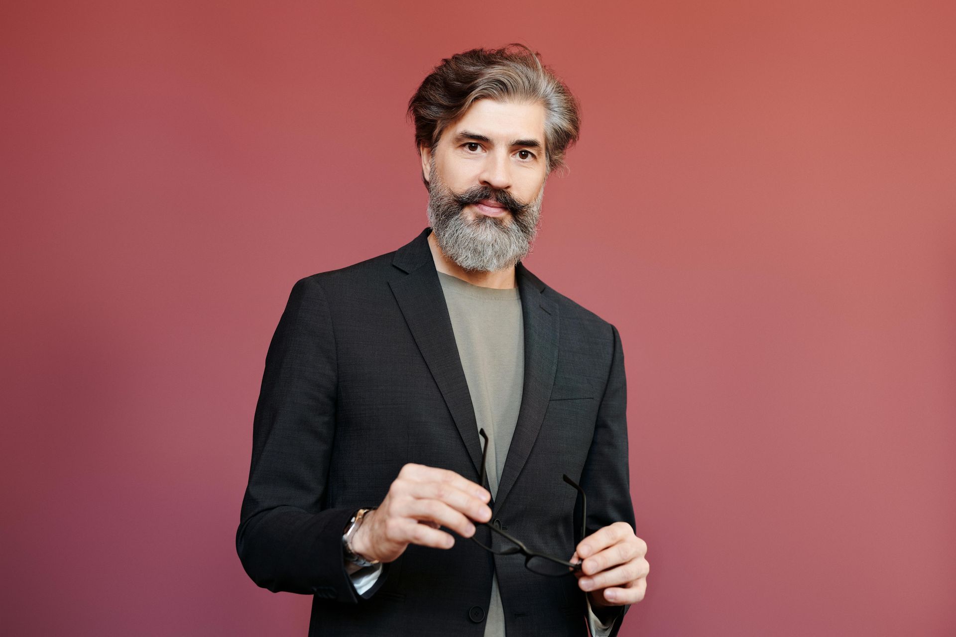 A man with a beard and mustache is wearing a suit and holding glasses.