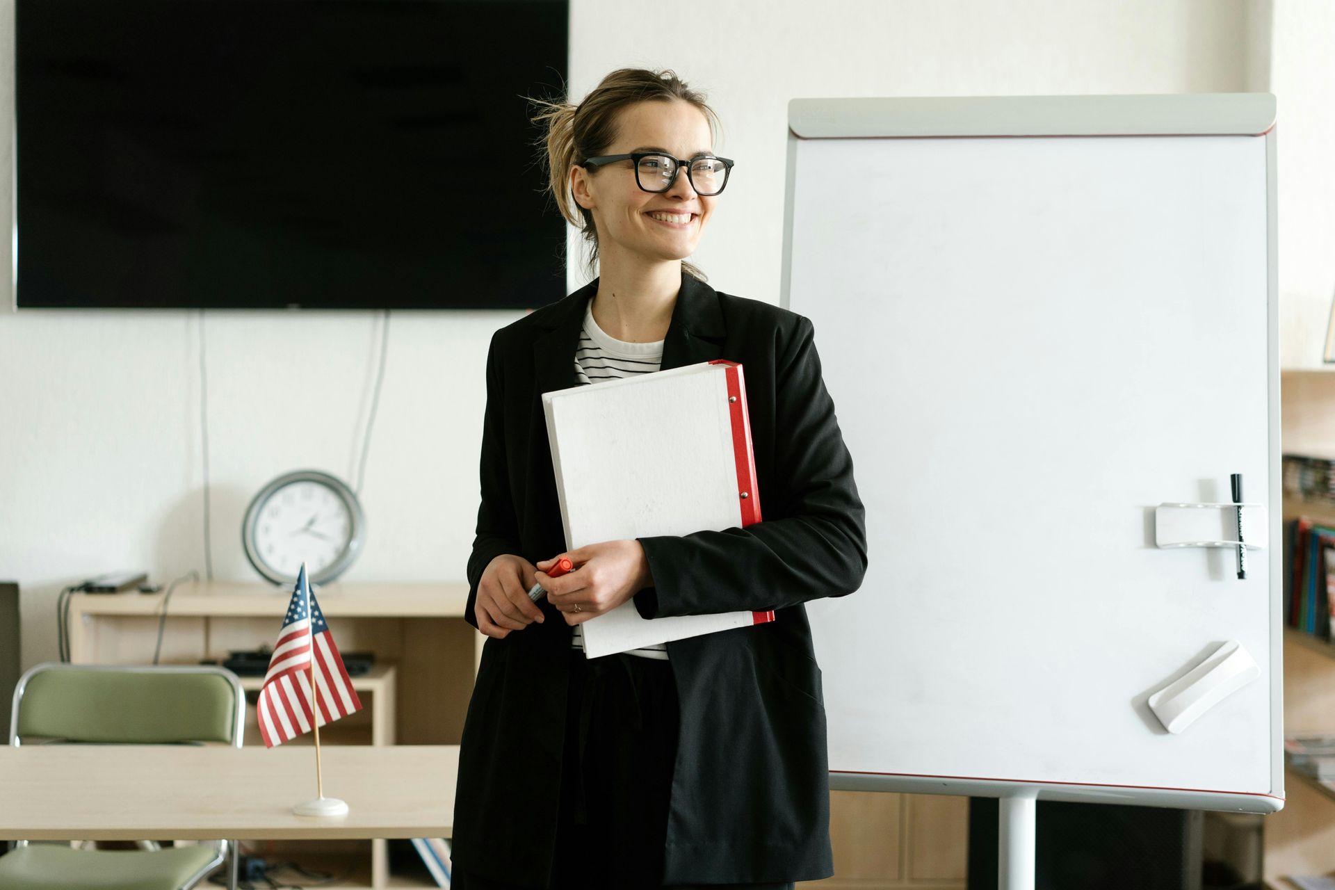 A woman is standing in a classroom holding a folder and smiling.