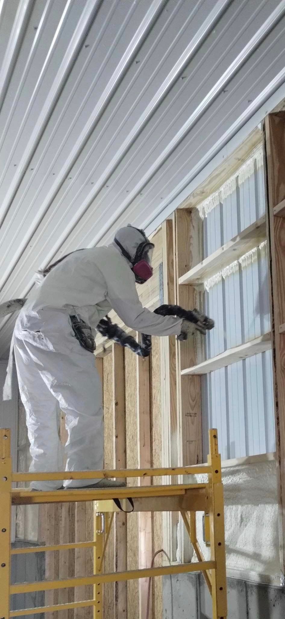 A man is spraying insulation on a wall while standing on a scaffolding.