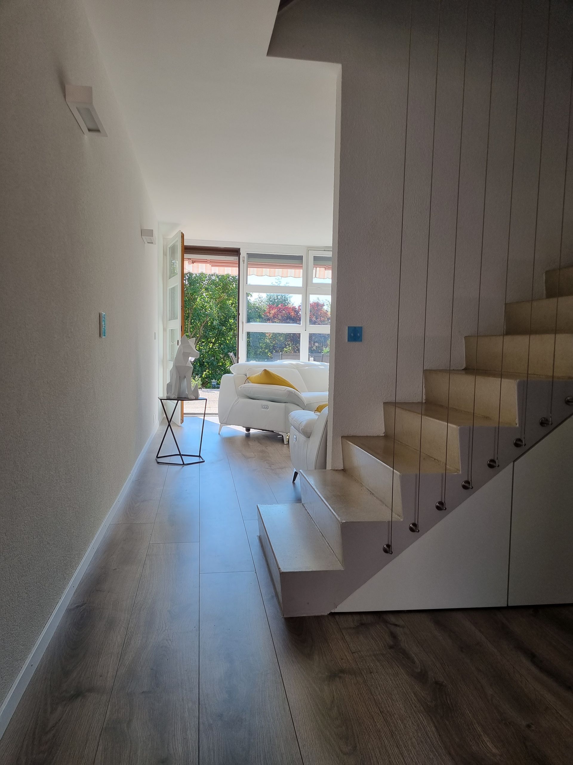 Couloir avec escalier menant à l'étage, vue sur un salon avec canapé blanc et plantes.
