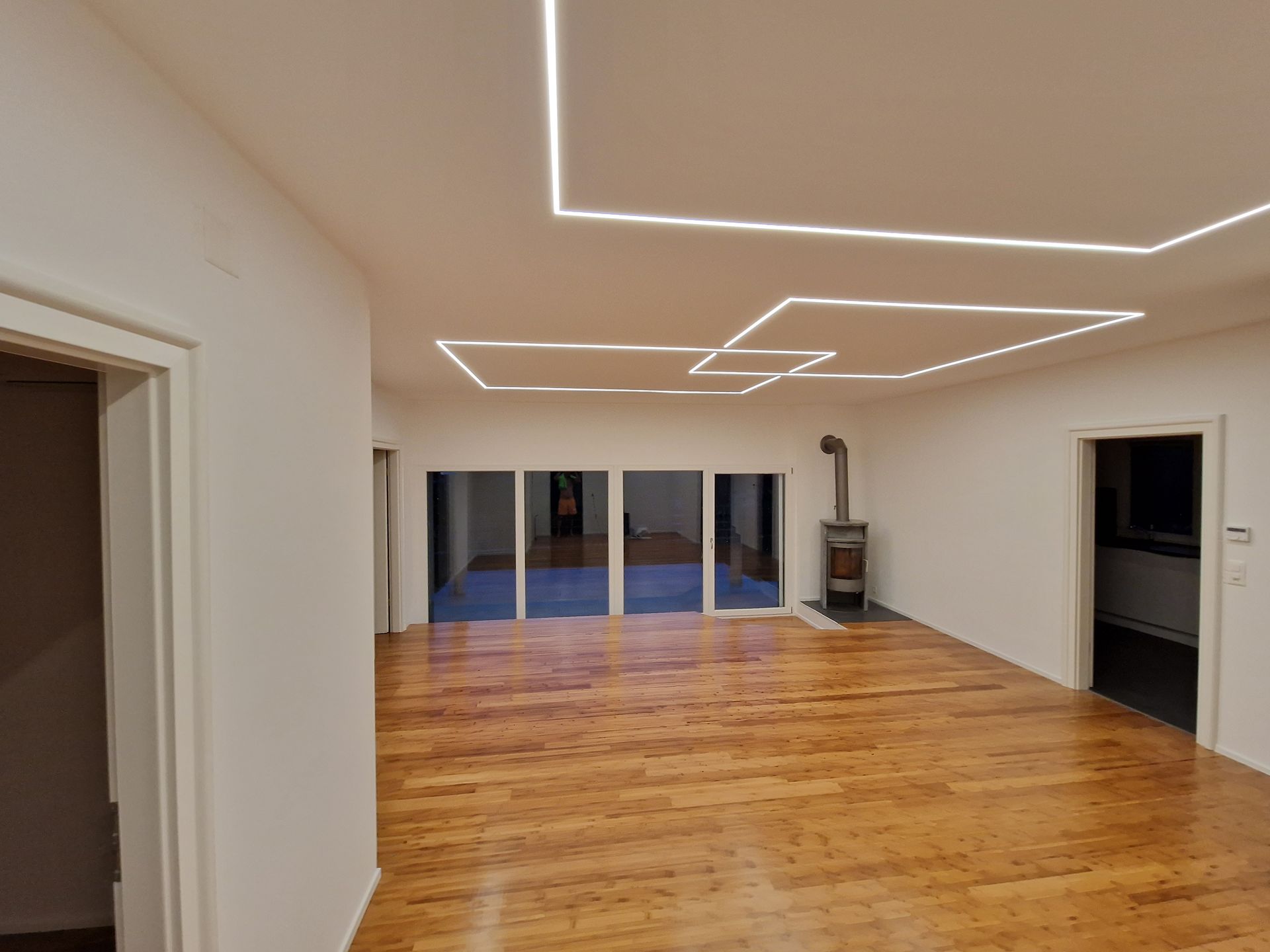 Pièce vide avec parquet, murs et plafond blancs avec éclairage linéaire, grandes fenêtres et cheminée.