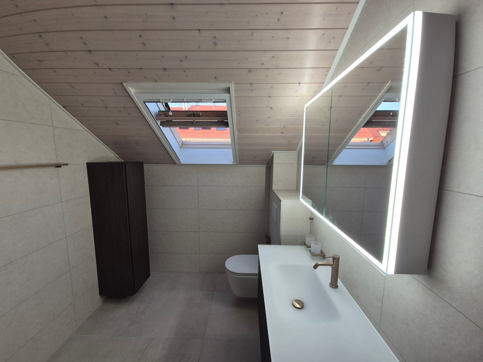 Salle de bains avec lavabo blanc, miroir éclairé, toilettes et plafond en bois avec puits de lumière.