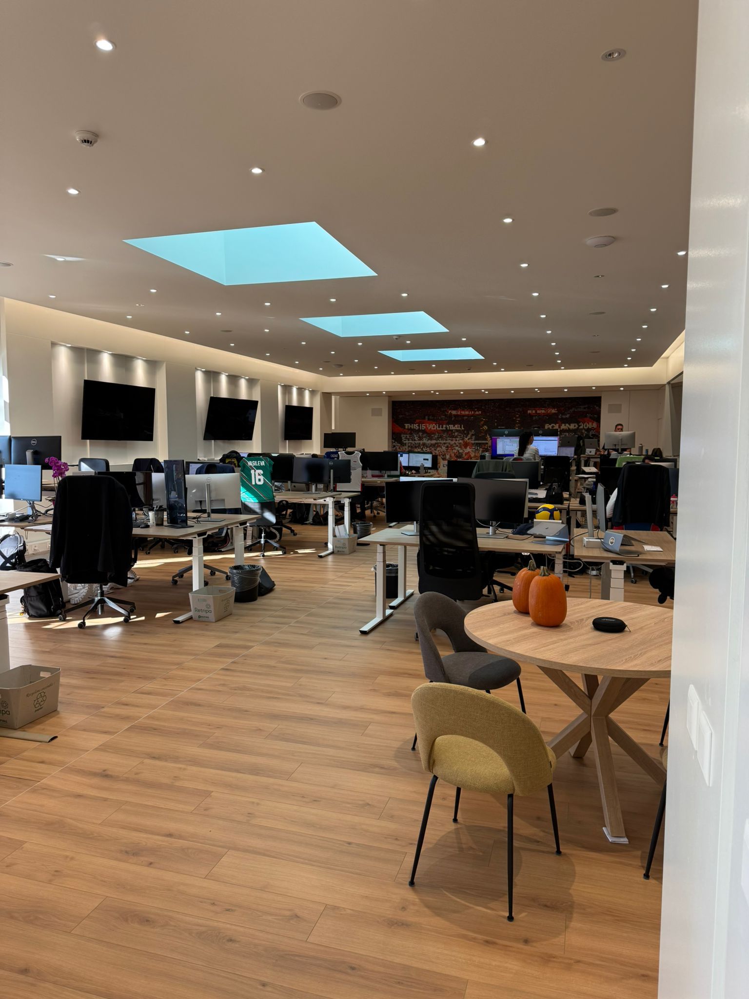 Un bureau à aire ouverte avec bureaux, ordinateurs et espaces de détente. La lumière naturelle pénètre par des puits de lumière.